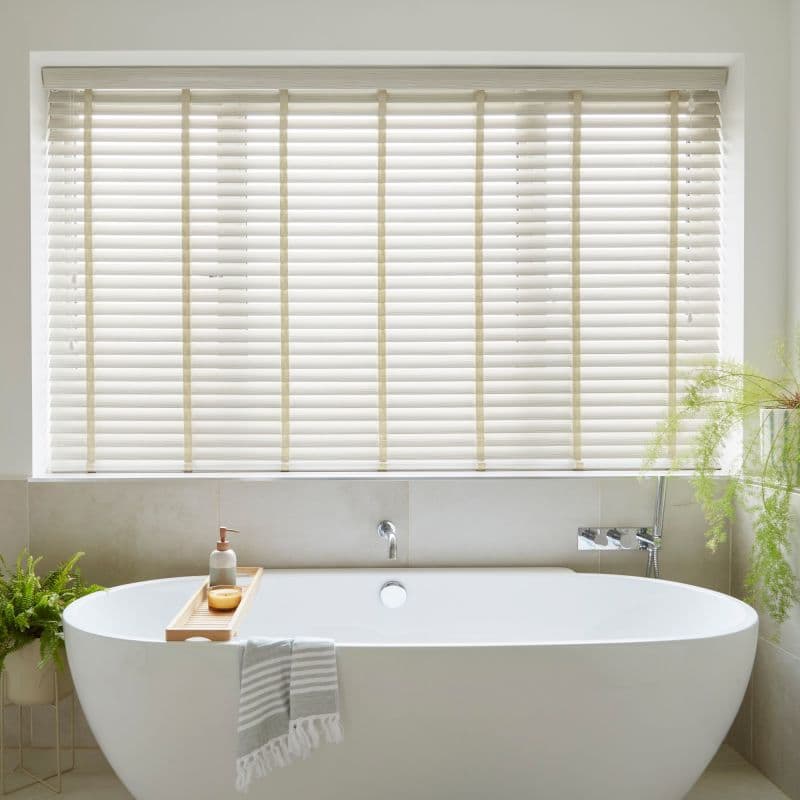 Cream horizontal slatted blinds with vertical ladder tapes cover a recessed window, closed and softly filtering daylight, above a freestanding white bathtub in a neutral, plant‑accented bathroom.