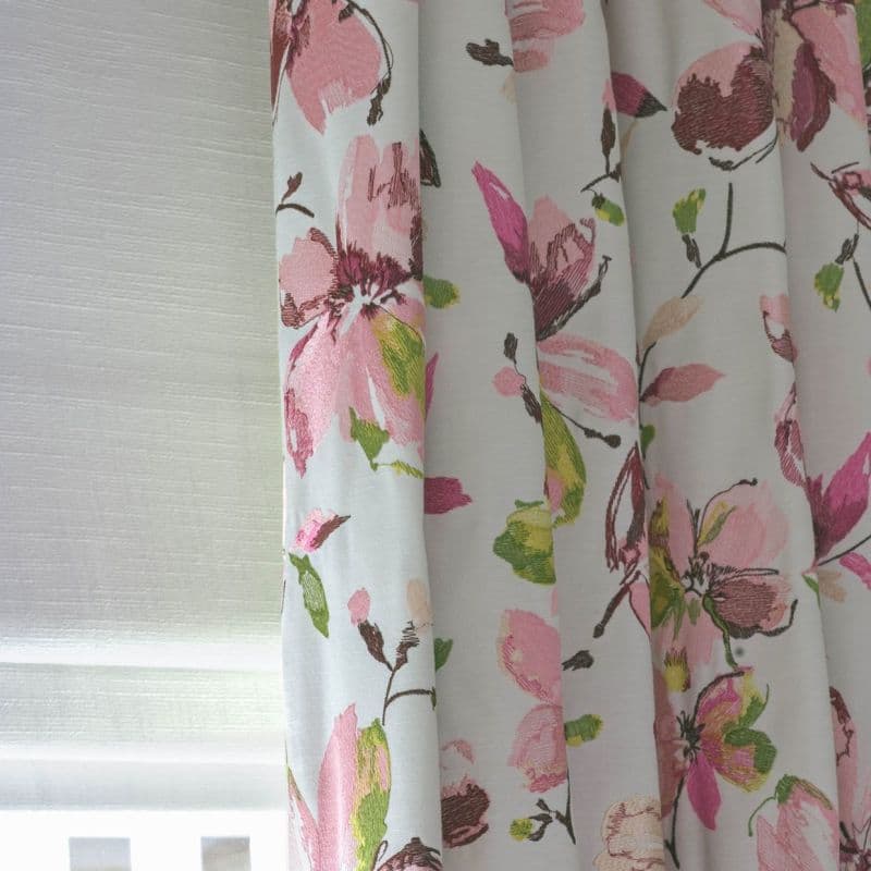 Floral fabric curtain — pleated and drawn at right with large pink and green blossoms — partially covering a lowered white roller blind, filtering soft daylight in a neutral-walled interior.