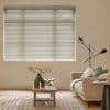 Horizontal white Venetian blinds cover a wide window, closed and softly filtering daylight; living room with beige sofa, wooden coffee table, sculptural vase and open book, patterned rug, floor lamp.