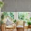Grey fabric Roman blinds, lowered with soft folds, partially filtering bright daylight through a narrow lower gap; sunlit sitting area with wicker chairs, patterned cushions, potted plants and garden view.