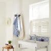 White café-style plantation shutters covering the lower window, slatted panels slightly ajar, filtering sunlight into a bright bathroom with a white wall-mounted sink, patterned tile backsplash, hanging towels.