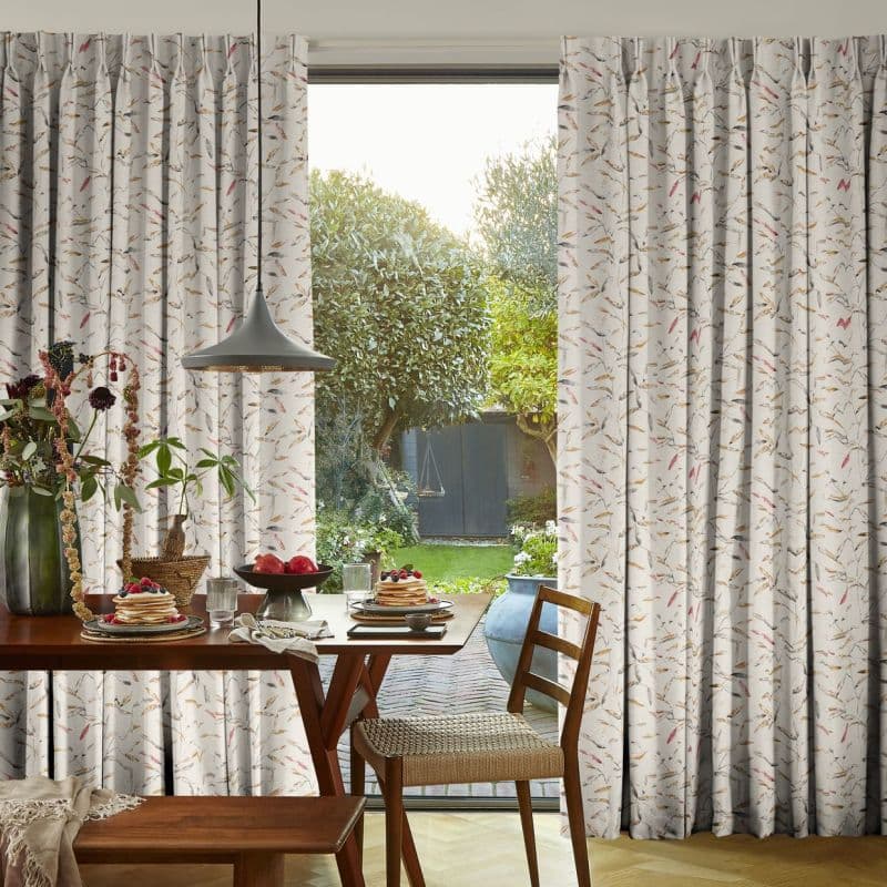 Patterned pleated curtains (light fabric with small leaf/feather print) framing a sliding glass door, parted to admit soft daylight; dining table set for breakfast with chairs and a lush garden view beyond.