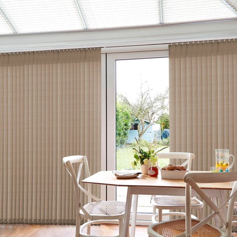 Beige fabric vertical blinds cover a sliding glass door, partially parted at center to reveal the outdoors, filtering light over a small dining table with white chairs and wooden floor.