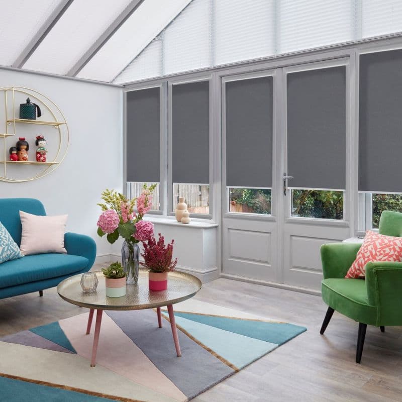 Grey roller blinds lowered over tall French doors and windows, leaving lower panes exposed and blocking light; in a bright conservatory-style living room with glass roof and colorful seating.