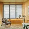 Horizontal slatted blinds covering three tall windows, semi-closed with alternating sheer and opaque bands — filtering soft daylight — warm wood-paneled sitting area with armchair, side table, potted plant, ambient under-cabinet lighting.