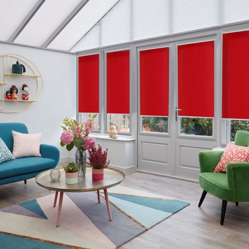 Red roller blinds lowered over four tall windows and a French door, covering upper panes while leaving lower panels exposed; blocking light in a bright conservatory with seating and flowers.