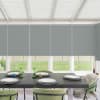 Dining table set with four place settings and green glassware sits beneath lowered grey roller blinds in a bright sunroom, green upholstered chairs and a grassy backyard visible.
