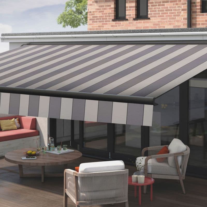 Striped retractable awning (diagonal grey and beige panels, scalloped valance) extended over sliding glass doors, shading a sunlit wood-deck patio furnished with cushioned chairs, a low table, and brick exterior.