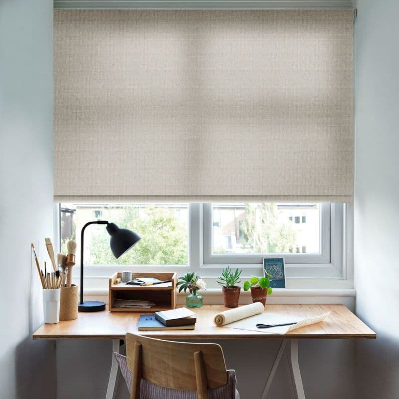 Beige fabric roller blind, fully lowered across two-pane window, softly diffusing daylight; above a wooden desk with lamp, brushes, notebooks and potted plants in a bright home workspace.
