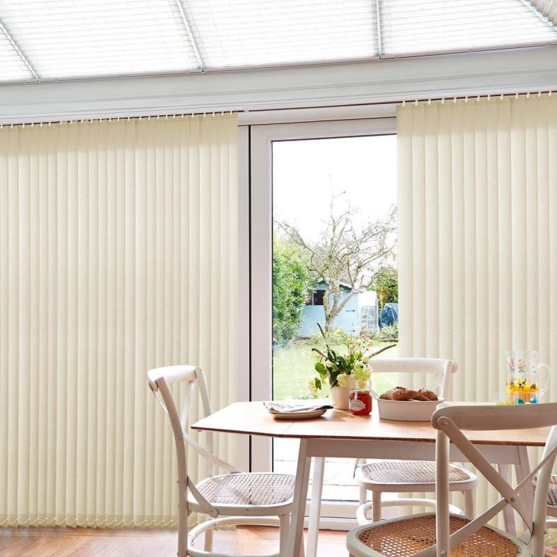 Cream vertical blinds covering a sliding glass door, panels mostly closed with a central gap revealing a garden; casting soft daylight into a dining area with wooden table and white chairs.