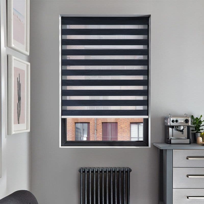 Black-and-gray zebra roller blind with alternating opaque and sheer horizontal bands covers the window, filtering light while a lower gap reveals a brick building; gray kitchen corner, espresso machine and radiator.