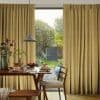 Floor-length pleated golden-yellow curtains with subtle texture, parted at center; filtering soft daylight and framing a glass door from a cozy dining area onto a sunlit, leafy backyard.