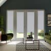 Glass French doors with patterned grey roller blinds lowered over panes, framing a sunny garden; situated within a living room containing green velvet armchairs, a glass coffee table, and a chevron rug.