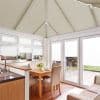 Cream pleated (honeycomb) blinds fitted across the pitched conservatory roof panels and over windows and French doors, closed and diffusing daylight; bright dining-kitchen conservatory with wooden table, beige chairs, patio view.