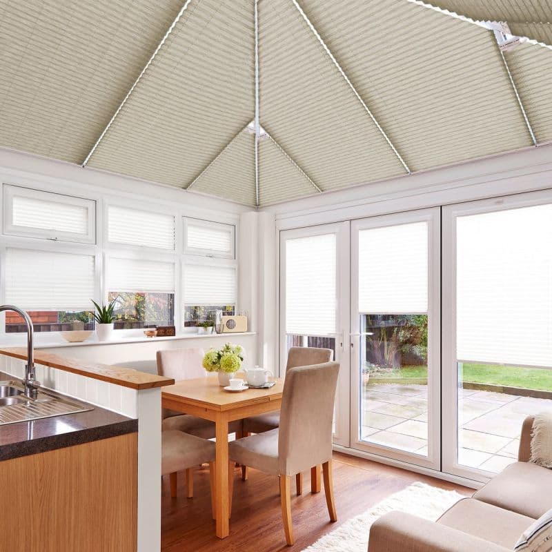 Cream pleated (honeycomb) blinds fitted across the pitched conservatory roof panels and over windows and French doors, closed and diffusing daylight; bright dining-kitchen conservatory with wooden table, beige chairs, patio view.