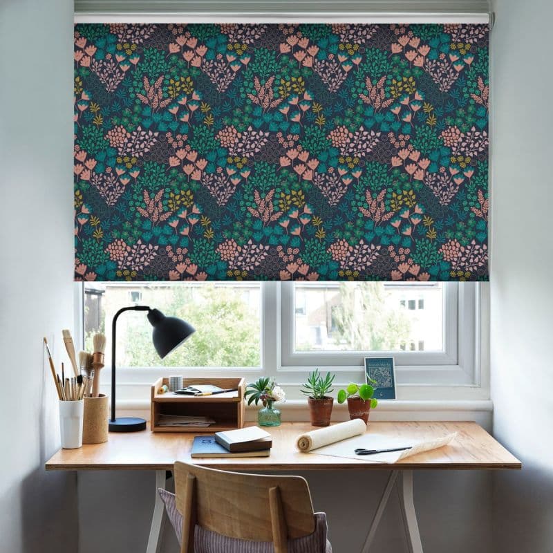 Roller blind with navy floral print, lowered across the window, filtering daylight. Tidy home workspace below — wooden desk with lamp, brushes and small potted plants, bright view outside.