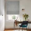 Grey textured Roman blind with vertical wave pattern; lowered to cover the window and filter daylight; above a wooden desk with teal chair, vase of flowers, mirror and coastal print.