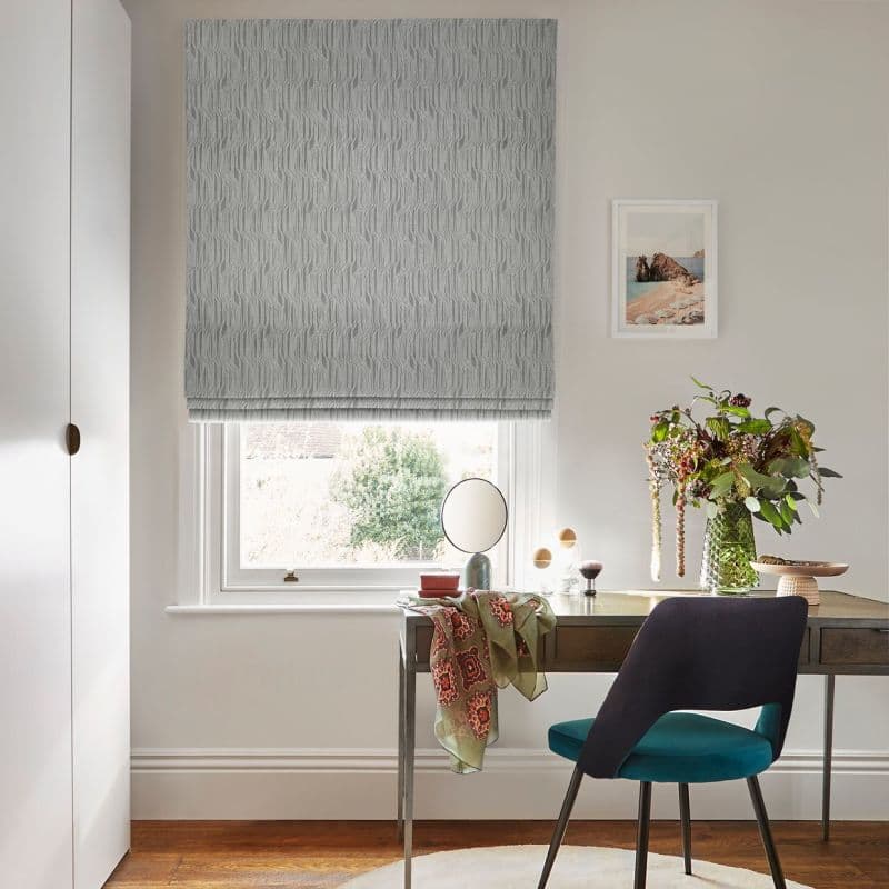 Grey textured Roman blind with vertical wave pattern; lowered to cover the window and filter daylight; above a wooden desk with teal chair, vase of flowers, mirror and coastal print.