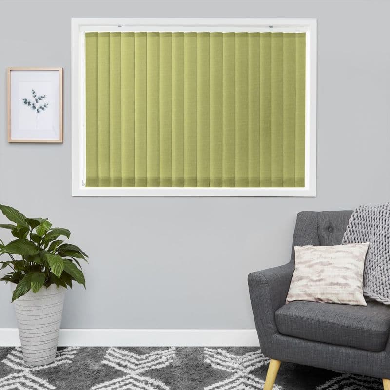 Vertical pale olive-green fabric blinds, closed inside a white-framed window with evenly spaced slats gently filtering daylight, set in a grey living room with a plant, armchair, and patterned rug.
