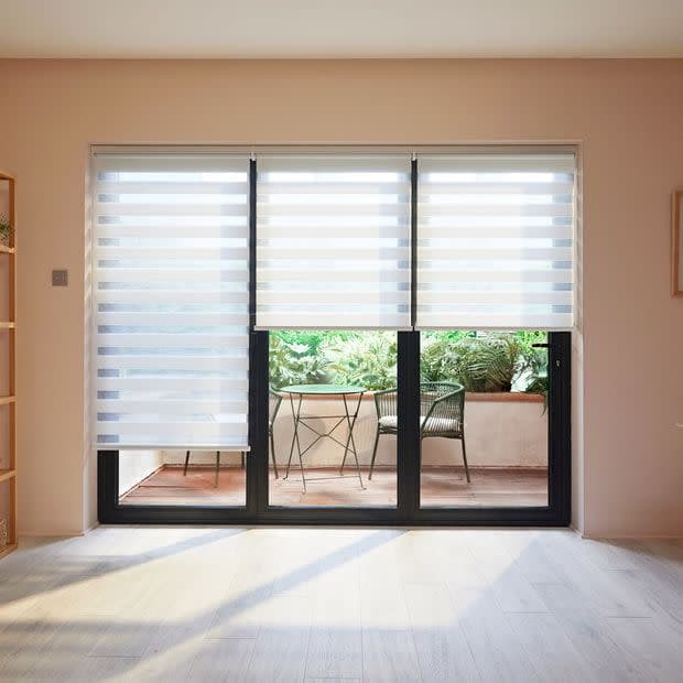 Three striped dual‑layer roller blinds over sliding glass doors, alternating sheer and opaque bands; partially lowered, filtering daylight onto pale wood‑floored room overlooking small balcony with table, chairs and greenery.