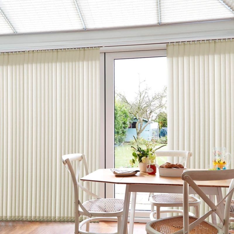 Cream vertical blinds cover a sliding glass door, mostly closed with a narrow central gap letting daylight through; a dining nook with wooden table, chairs, flowers, pastries and backyard view.