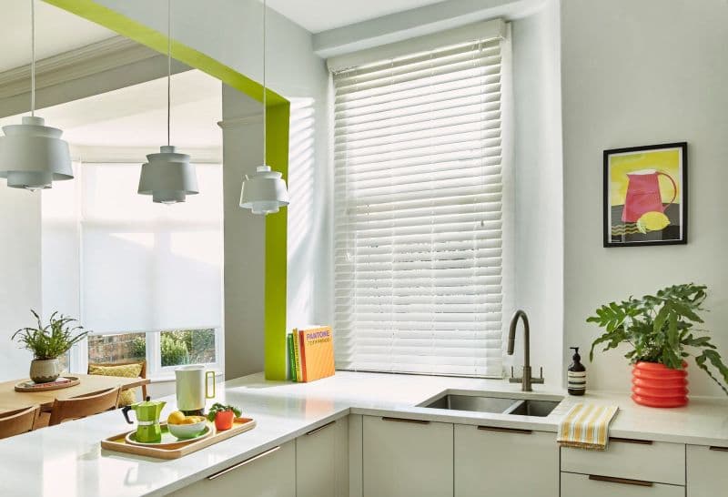 White horizontal blinds covering a tall kitchen window, closed and slightly tilted to filter sunlight and cast striped shadows; modern, bright kitchen with marble countertops, sink, pendant lights, and plants.
Text found: PANTONE