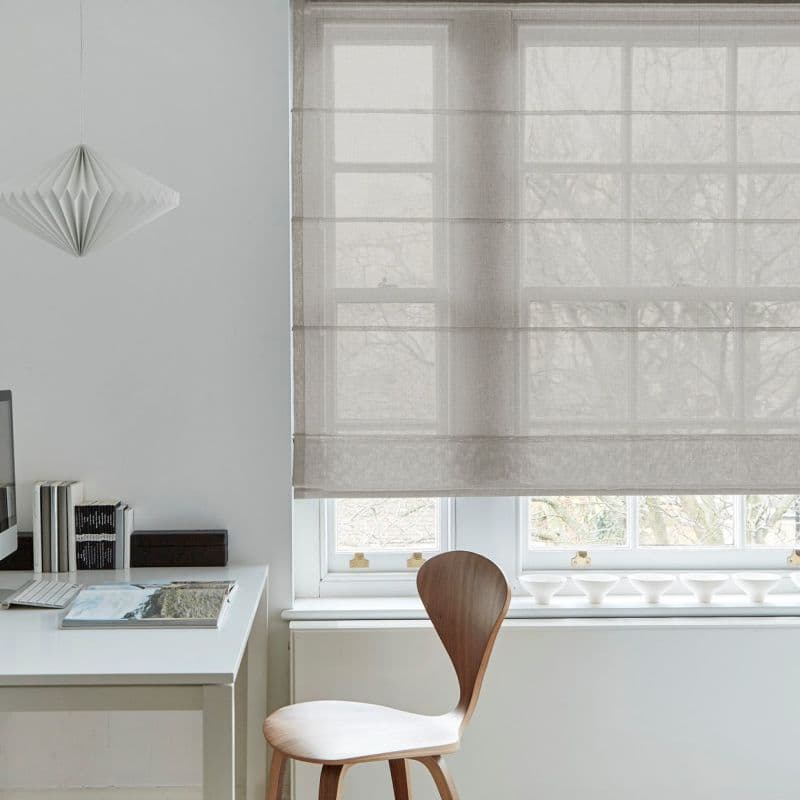 Beige semi-sheer Roman shade lowered across a multi‑pane window, horizontal folds visible; filtering diffuse daylight; minimal home office with white desk, wooden chair, pendant lamp and faint tree silhouettes outside.