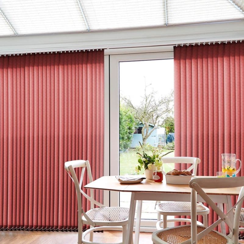 Coral vertical blinds with narrow slats, mounted over a patio glass door, partially drawn to reveal the garden; daylight filters into a dining area with table, chairs, vase and pastries.