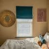 Teal Roman shade mounted above a window, partially lowered over white horizontal blinds, filtering daylight; bedroom wall with sunburst mirror left, abstract line-art print right, sunlit bed below.