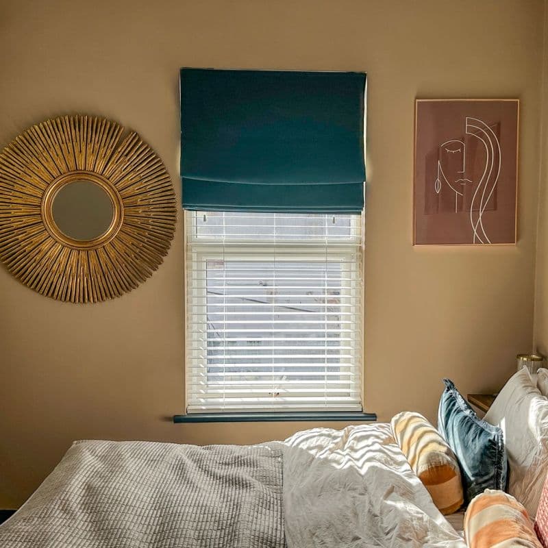 Teal Roman shade mounted above a window, partially lowered over white horizontal blinds, filtering daylight; bedroom wall with sunburst mirror left, abstract line-art print right, sunlit bed below.