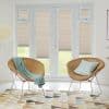 Beige pleated blinds on French doors and side panels, mostly lowered and softly filtering daylight; set in a bright living room with two rattan chairs, pastel cushions, and geometric rug.