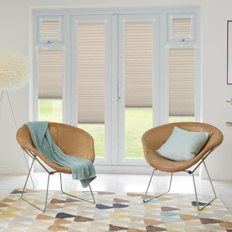 Beige pleated blinds on French doors and side panels, mostly lowered and softly filtering daylight; set in a bright living room with two rattan chairs, pastel cushions, and geometric rug.