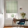 Striped Roman blind (green, grey, white vertical stripes) lowered, filtering soft daylight through a sash window; home desk with teal chair, vase of flowers, scarf-draped tabletop and framed print.