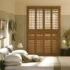 Wooden full-height plantation shutters covering a tall window—four hinged panels with louvered slats—closed and filtering warm daylight into a neutrally toned bedroom with bed, lamps, and wall decor.