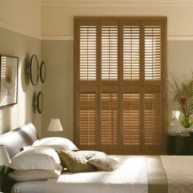 Wooden full-height plantation shutters covering a tall window—four hinged panels with louvered slats—closed and filtering warm daylight into a neutrally toned bedroom with bed, lamps, and wall decor.