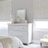 Patterned fabric Roman shades, lowered and filtering daylight through pale geometric motifs; positioned over two windows above a white distressed three-drawer dresser — neutral bedroom with mirror, vases, soft lighting.