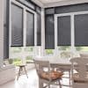 Dark horizontal slatted blinds covering tall bay windows, mostly lowered to mid-height. They filter and block light. Bright sunroom/dining area with white table and chairs, garden visible through lower panes.