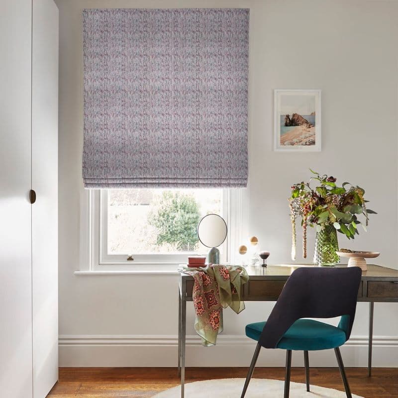 A speckled fabric Roman blind, lowered over a sash window, gently filtering daylight; in a bright home office with a desk, teal chair, small round mirror, vase of foliage and framed coastal print.