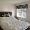 Floral Roman valance above white slatted blinds, blinds tilted to filter daylight; bedroom window beside a double bed with textured white coverlet, floral pillows, neutral walls and wooden bedside table.