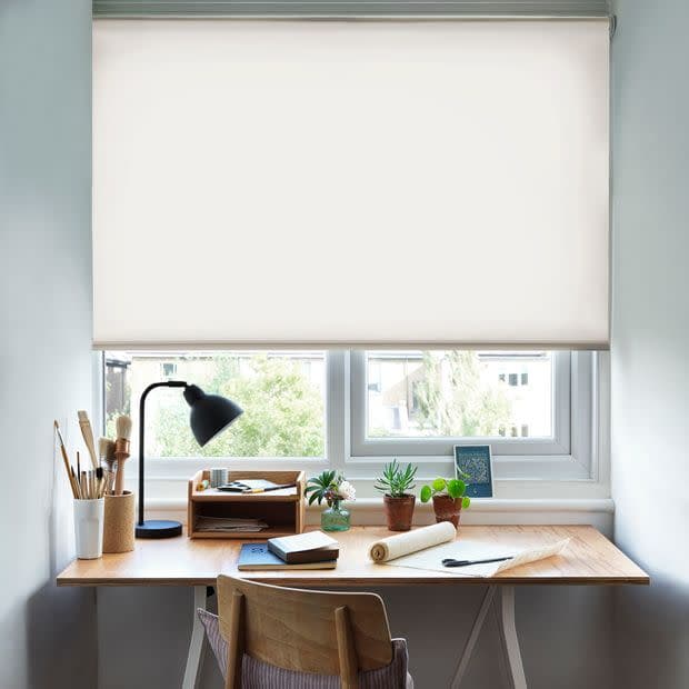 White roller blind, lowered across the window, diffusing bright daylight; home study desk beneath with wooden surface, lamp, potted succulents, notebooks and brushes, well-lit workspace.