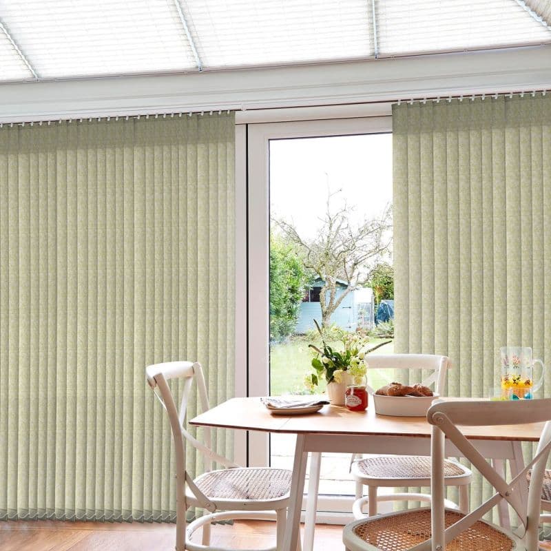 Vertical pale-green fabric vertical blinds covering sliding glass doors, mostly closed with a central opening revealing daylight; small dining table with white chairs, vase and breakfast items in sunlit conservatory overlooking garden.