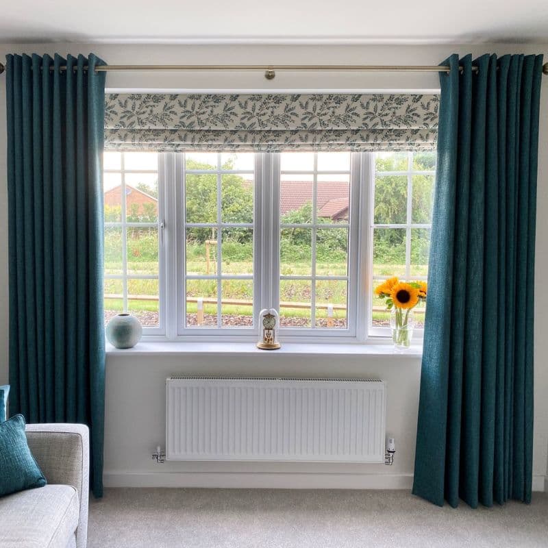 Teal grommet curtains pulled to sides, with patterned Roman blind lowered across top, framing and filtering daylight in a bright living room; windowsill sunflowers and clock, radiator below, garden beyond.