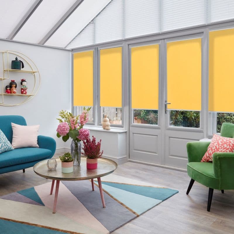 Yellow roller blinds, lowered across four tall glass doors and windows, blocking light; contemporary conservatory-style living room with pastel furnishings, geometric rug, plants, and soft daylight from a translucent roof.