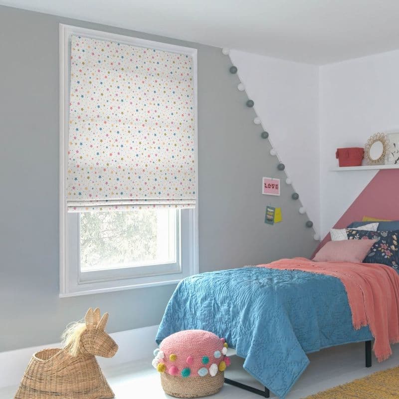 Star-patterned fabric roman blind fully lowered over the window, white with small multicolored stars, filtering soft daylight; in a girl's bedroom with gray/pink walls, blue quilt, string lights, wicker toy and pom‑pom pouf. LOVE