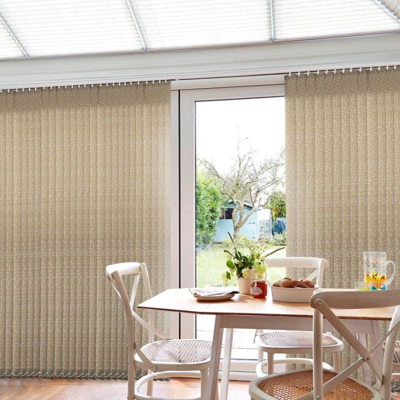 Beige patterned vertical blinds, floor-to-ceiling and mostly closed across a sliding glass door, slightly parted to a garden; filtering daylight into a small dining area with wooden table, chairs, flowers.