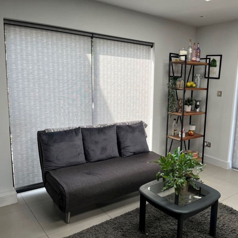Light-gray roller blinds, fully lowered over sliding glass doors with subtle woven texture, filtering soft daylight; a charcoal sofa with a glass coffee table and shelving in a living room.