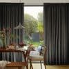 Floor-length dark gray pleated curtains, gathered to sides, pulled open at center, framing a dining table with lamp and chairs, opening onto a sunlit backyard with trees and gate.