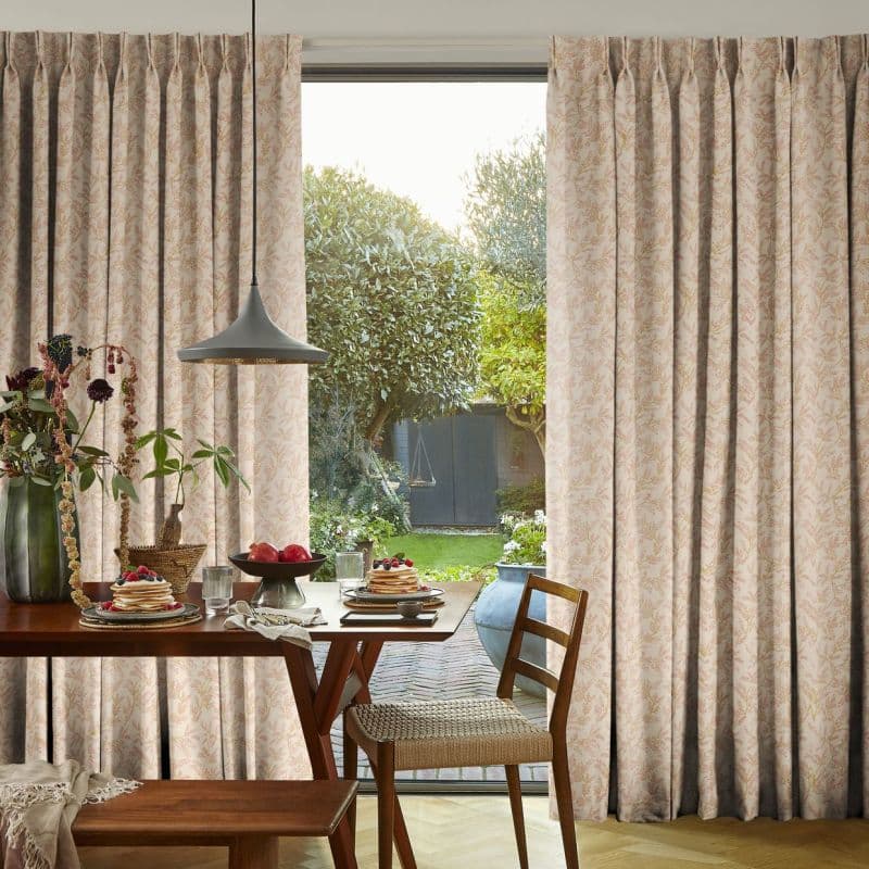 Patterned, pleated full-length curtains, pulled slightly open to reveal a central glass door; hanging and filtering soft daylight. Wooden dining table set for breakfast with view of a green garden beyond.
