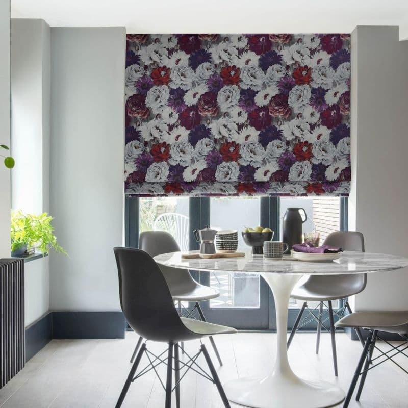 Floral Roman blind, lowered over patio doors with dense white, purple and red blooms; filtering soft daylight into a modern dining nook with a white tulip table, grey chairs, plant.