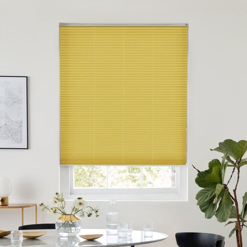 Yellow pleated blind lowered nearly to the sill, filtering light; set in a bright white dining room with a round table, glassware, framed art, and a potted fiddle-leaf fig.